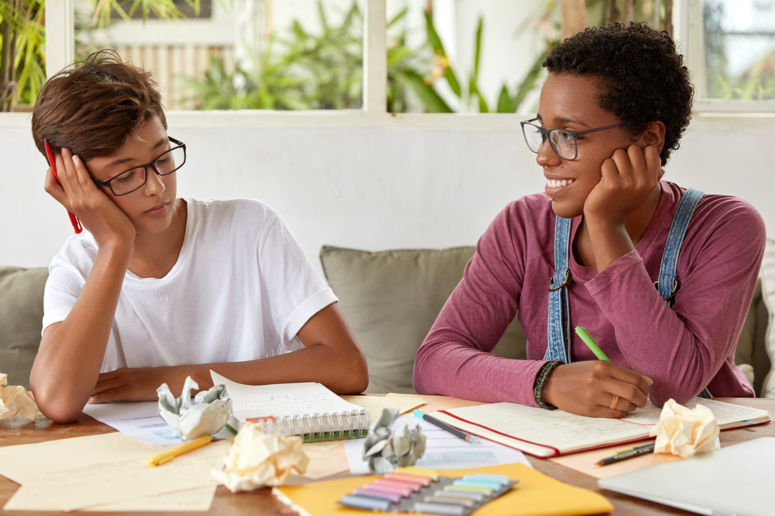 Diverse girl and boy meet to do homework, make notes in notebook, give advice to each other, talk about common project, share ideas, sit next to each other at table, cooperate for making course work